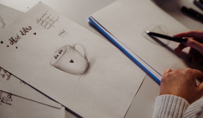 Proces projektowania kubków w pracowni marki Nadzwyczajnie.pl - zbliżenie na szkic kubka z sercem, ręce projektantki z ołówkiem oraz zapiski koncepcyjne na białym papierze