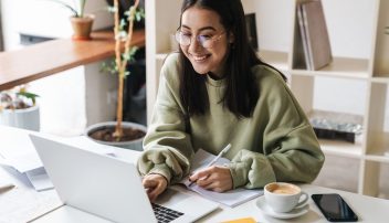 Uśmiechnięta młoda kobieta w okularach i luźnej bluzie pracuje przy laptopie, notując w zeszycie – tworzy strategię marketingu afiliacyjnego przy filiżance kawy w nowoczesnym, przytulnym wnętrzu