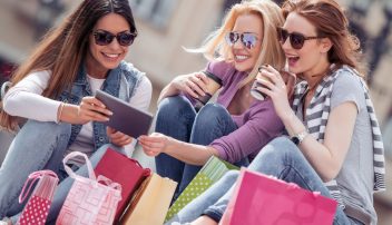 Grupa młodych kobiet z zakupami i kawą przegląda tablet – analizują możliwości sprzedaży detalicznej w e-commerce.”